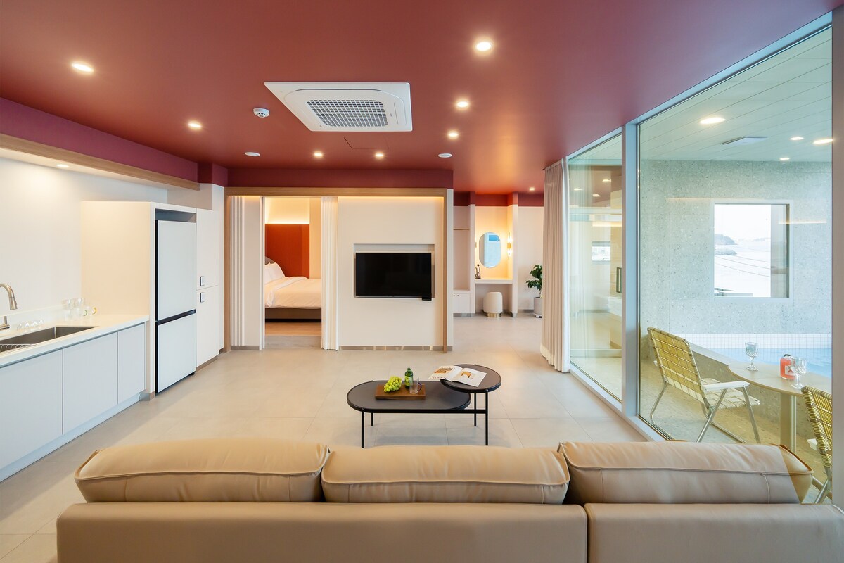 An open living space features a comfortable sofa facing a wall-mounted television. A sleek kitchen area includes a refrigerator and modern cabinetry. Large glass doors offer views to the ocean and allow natural light to fill the room, connecting indoor and outdoor environments.