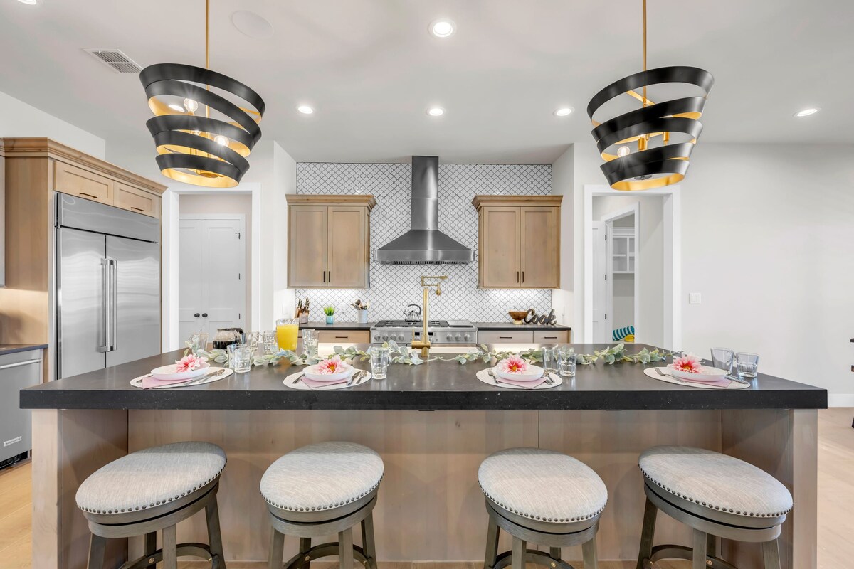 A modern kitchen features a large island with four bar stools, complemented by elegant pendant lights above. The sleek cabinetry in warm wood tones and a stylish backsplash visually enhance the space. Breakfast items are artfully arranged, inviting guests to enjoy a meal together.