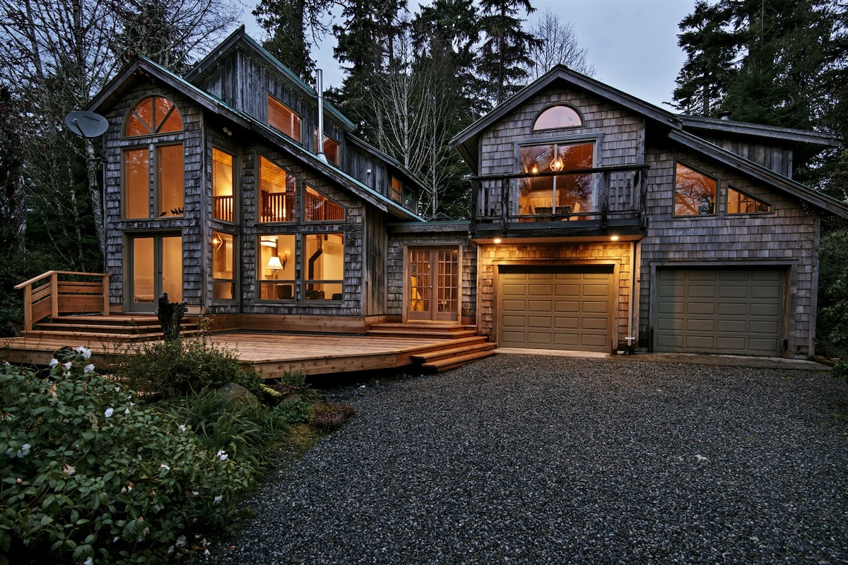 The exterior of a two-story house is showcased in natural surroundings, with wood paneling and large windows reflecting the evening light. A spacious wooden deck extends from the front, while a gravel pathway leads to the twin garage doors. The vibrant greenery is accented by soft outdoor lighting.