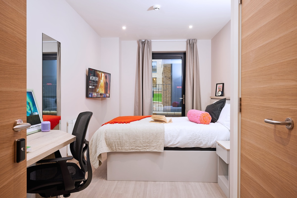 A comfortable bedroom is seen featuring a spacious bed with a light-colored blanket, accompanied by colorful pillows. A wall-mounted Smart TV is positioned for entertainment. Natural light filters through curtains, illuminating a workspace with a desk and chair. A door is visible, leading to an outside area.