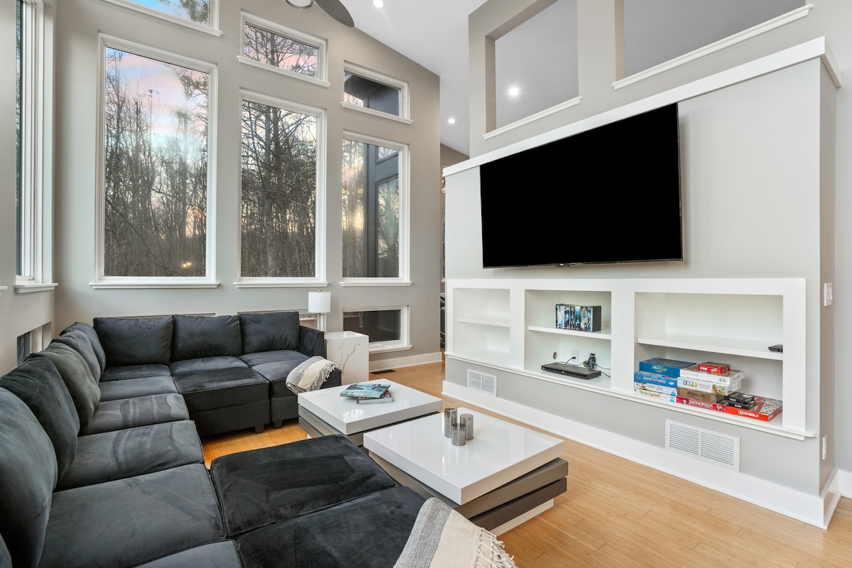 A contemporary living area features a large sectional sofa arranged around a low coffee table. A wall-mounted television is displayed above a sleek media console, and large windows allow natural light to fill the space, offering views of the surrounding trees.