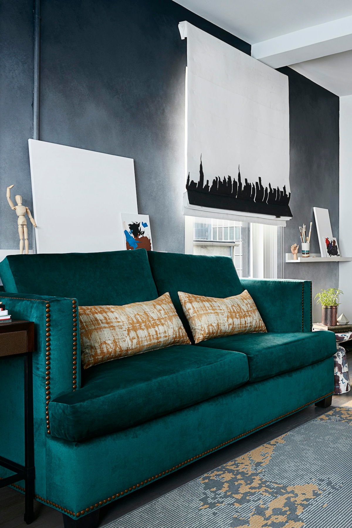 A plush teal sofa with decorative cushions is positioned against a textured dark wall. Nearby, a large window is partially covered by a rolling shade featuring a skyline silhouette. Various pieces of art and decor are displayed on adjacent shelves.