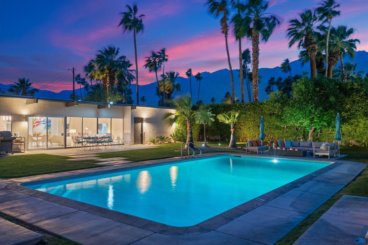 An illuminated pool is framed by lush greenery and palm trees, reflecting the vibrant colors of the sunset sky. The modern home features large glass doors, providing a view into the elegant interior. Comfortable outdoor seating is arranged nearby, enhancing the tranquil setting.