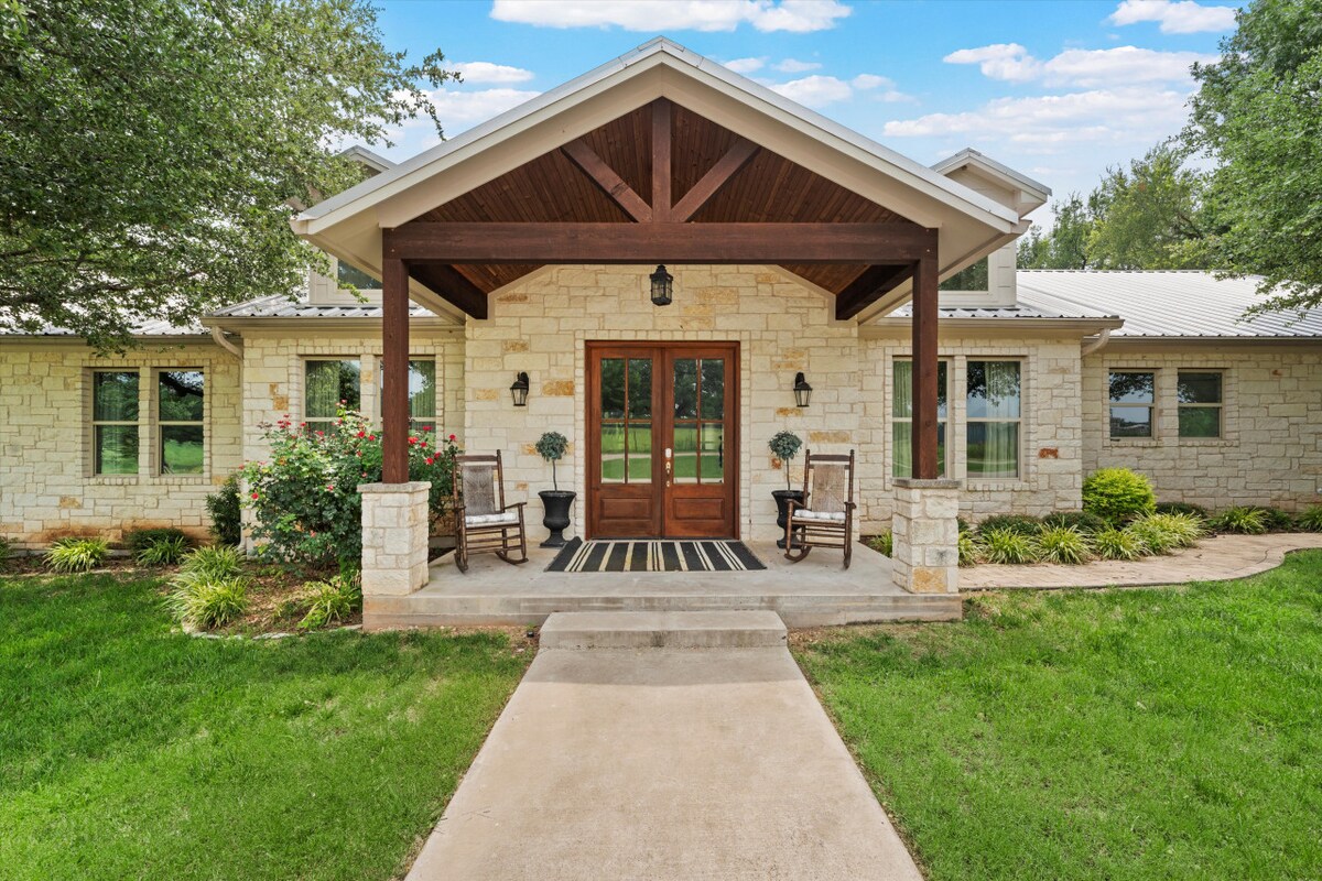The entrance features a covered porch with wooden beams, symmetrical rocking chairs, and well-maintained landscaping. A stone pathway leads to double doors flanked by outdoor sconces, while a grassy area surrounds the home, creating a warm and welcoming approach.