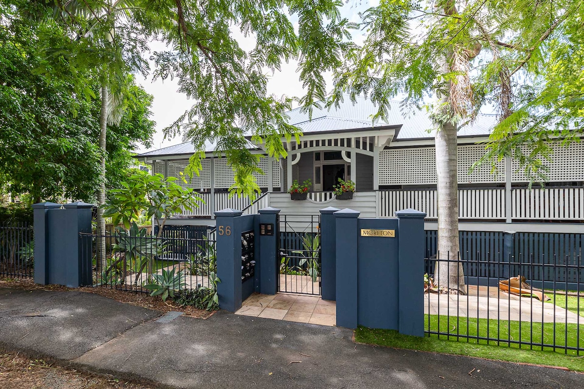 The entrance to a historic Queenslander home is framed by lush greenery and mature trees. A charming blue gate leads to a pathway with stone tiles, bordered by manicured lawns and shrubs. The elevated porch features decorative latticework, inviting guests into the serene environment.