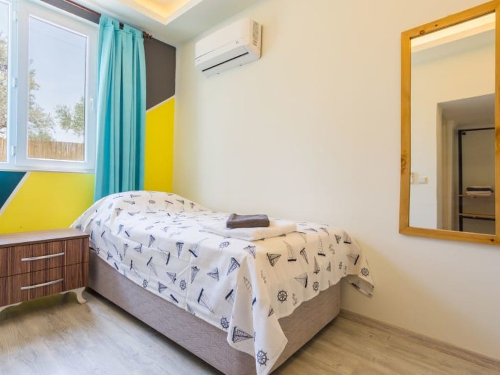A bright sleeping area is characterized by a spacious bed covered with a white patterned blanket. Natural light enters through a large window adorned with sheer turquoise curtains. A mirrored accent reflects the room's modern design, complemented by a wooden nightstand beside the bed.