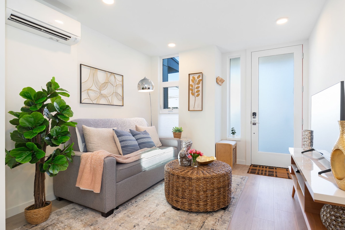 A cozy living room features a grey sofa adorned with light throw blankets. A circular woven coffee table sits in front of the sofa, complemented by decorative items. Natural light enters through a nearby window, enhancing the bright interior atmosphere.