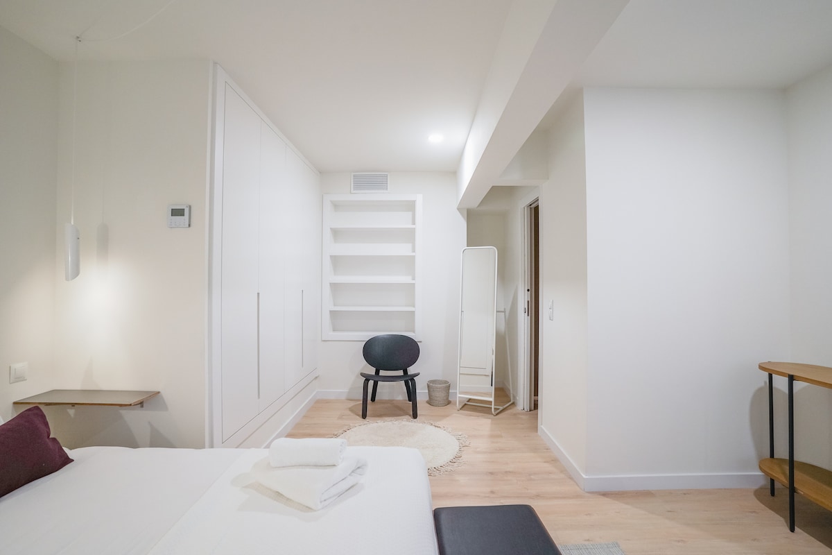 A bright bedroom space includes a spacious bed with fresh linens and two neatly folded towels. A minimalist shelving unit occupies one wall, while a round rug is positioned beneath a single chair. A door leads to an adjacent bathroom, enhancing the room's functionality.