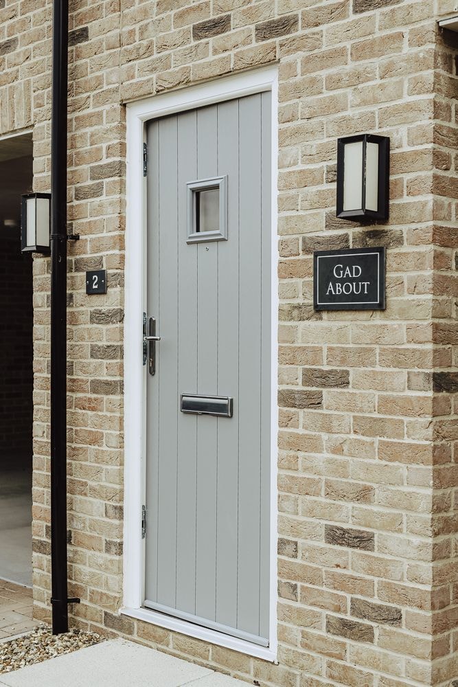 The entrance features a modern grey door set within a textured brick facade. A glass panel allows for light while maintaining privacy. The nameplate 'GAD ABOUT' is affixed beside the door, accompanied by two wall-mounted light fixtures that enhance visibility.