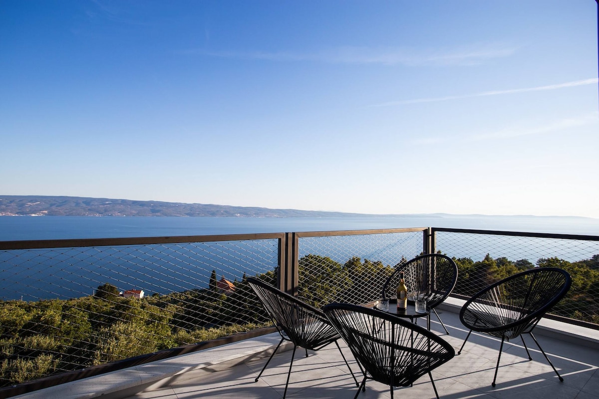 A balcony offers a sweeping view of the Adriatic Sea, with hills and coastline visible in the distance. Four black lounge chairs are positioned around a small table, inviting relaxation while enjoying the scenic surroundings and tranquil atmosphere.