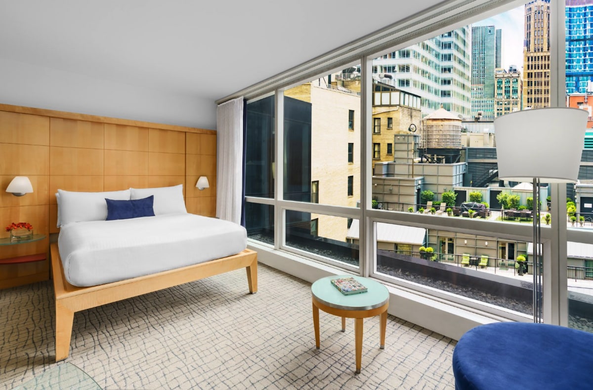 A spacious room features a queen bed with a light-colored duvet. Large windows offer a view of the cityscape, with greenery visible on nearby rooftops. A small round table and a blue armchair provide additional seating options. Modern lighting complements the wood-paneled wall.
