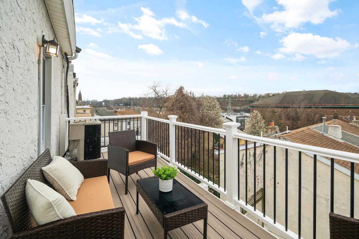 A sunlit deck features comfortable seating with neutral-colored cushions and a small table. A potted plant adds a touch of greenery, while expansive views of the surrounding area and distant buildings create an inviting outdoor space.