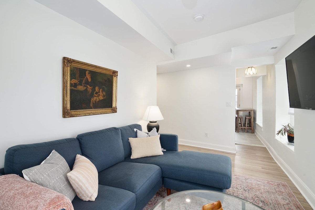 A cozy living room is furnished with a blue sectional sofa, accented by decorative throw pillows. A framed painting adorns the wall, while a glass coffee table sits in front. Soft lighting from a lamp creates a comfortable space, with an open view into the dining area.