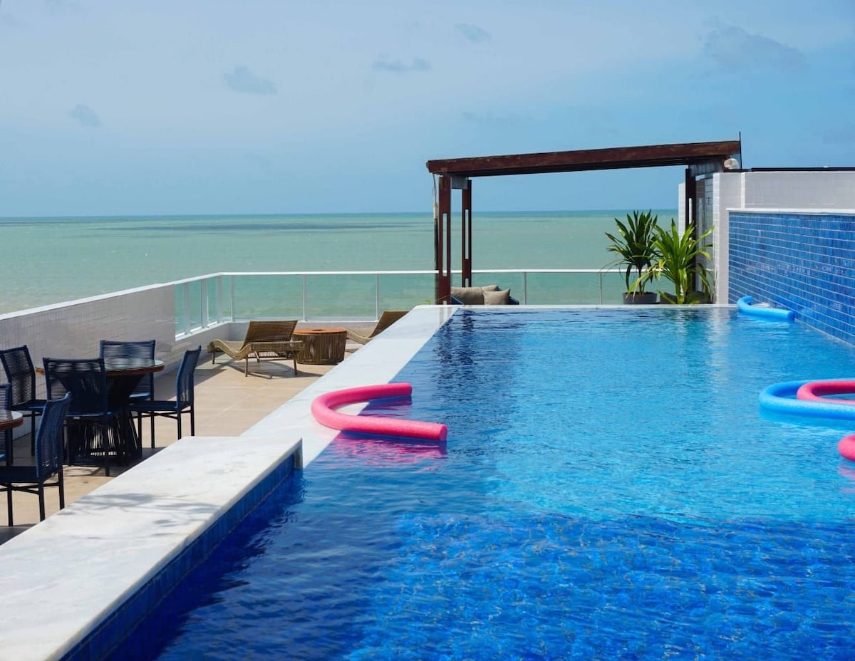 A panoramic pool area is presented with tranquil blue waters extending toward the horizon. Comfortable lounge chairs are arranged nearby, and vibrant pool floats are visible in the water. The area features modern furnishings and a view of the sea, creating a relaxing outdoor space.