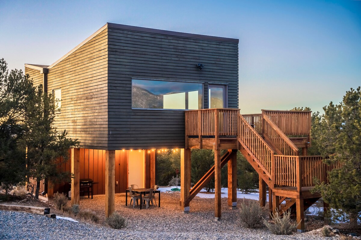 A modern eco-friendly home is elevated on stilts, showcasing large windows that invite natural light. A gravel driveway leads to an outdoor wooden staircase. The surrounding landscape features trees and open space, creating a serene setting at dusk.
