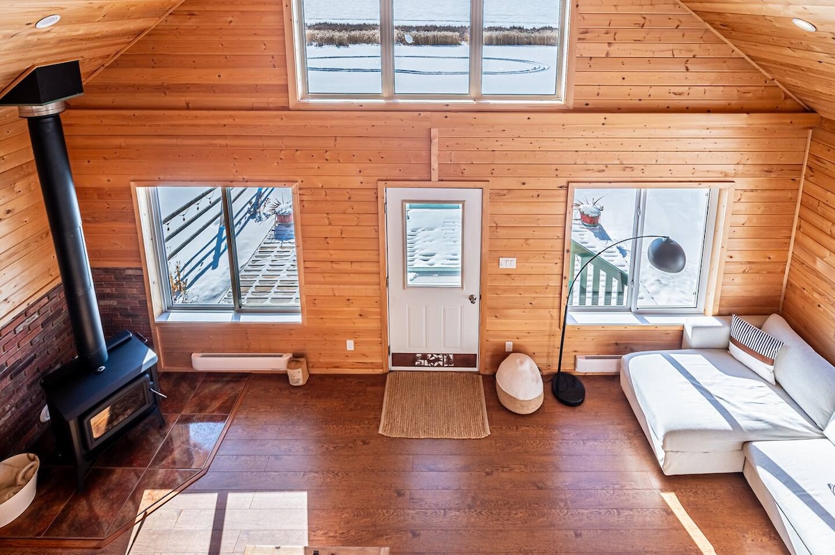 A bright interior space showcases rustic wooden walls and a high ceiling, complemented by large windows providing ample natural light. A door leads outside, while a comfortable sofa and a modern floor lamp create a cozy seating area. A wood stove is positioned for warmth.