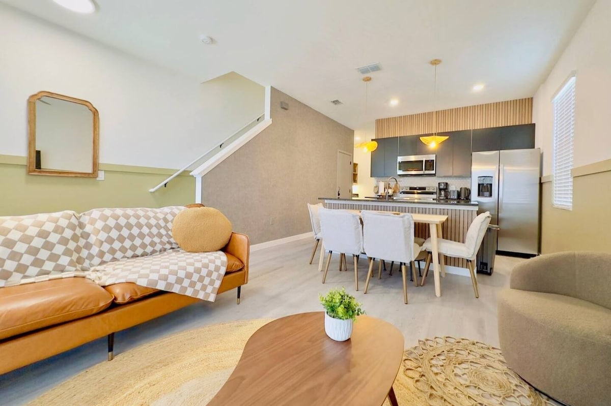A stylish living area features a combination of a patterned sofa and a large round coffee table. A spacious dining table with wooden chairs is visible in the background, accompanied by modern stainless steel appliances in the kitchen. Soft lighting fixtures create a warm ambiance.