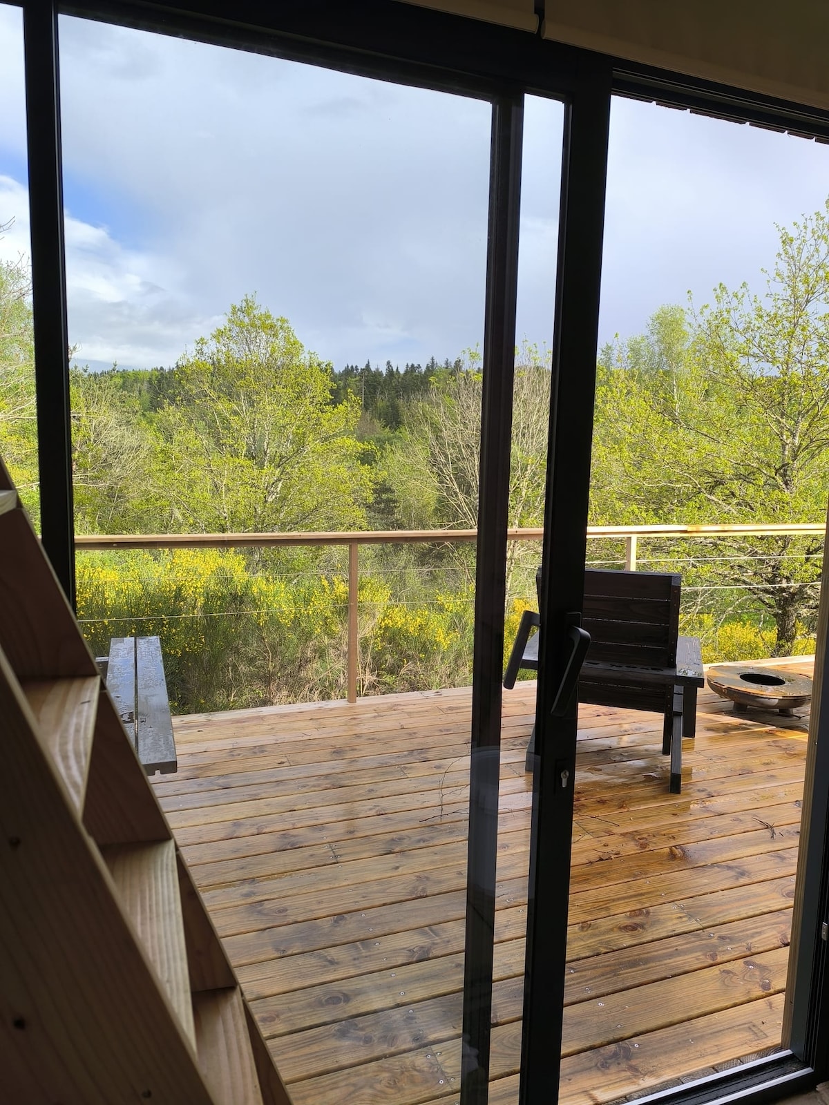 A wooden deck is visible through sliding glass doors, showcasing a natural landscape of lush green trees. A single chair and a fire pit are positioned on the deck, which features a warm wooden floor. The sky above is partly cloudy.