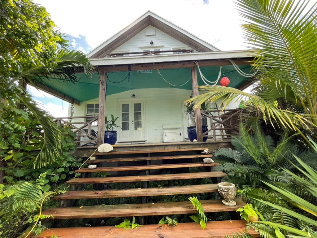 The entrance to the farmhouse features a set of wooden steps leading up to a covered porch, framed by lush tropical greenery. Potted plants are placed on either side of the doorway, and a decorative lantern adds a subtle touch of charm.