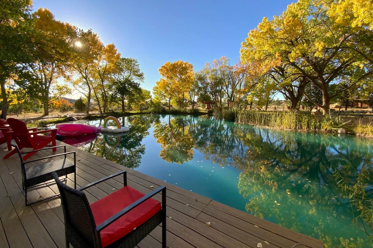 A peaceful pond is surrounded by vibrant autumn foliage, reflecting the colorful trees in the still water. Wooden decking features two red chairs, offering a serene spot to relax and enjoy the natural beauty. Sunlight filters through the trees, enhancing the tranquil setting.