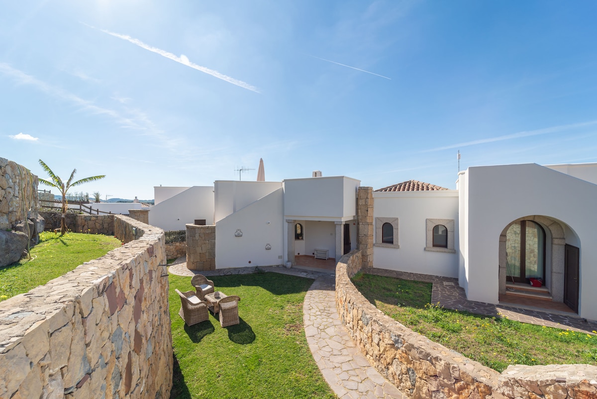 The villa's exterior is showcased with a curved pathway leading to the entrance. Surrounding stone walls enclose a green lawn, while outdoor seating is arranged for relaxation. Natural light bathes the area, enhancing the clean architectural lines and the welcoming ambiance of the space.