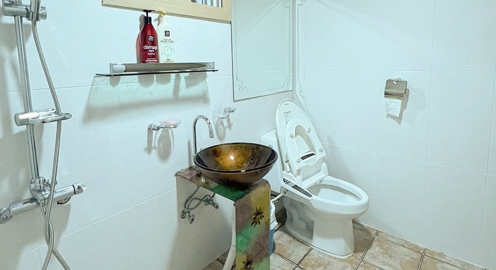 A modern bathroom is presented with clean white tiles and warm, earthy tones. A decorative sink crafted from a unique material is positioned on a sturdy stand. A stylish toilet and necessary toiletries are also visible, ensuring a functional and inviting space.