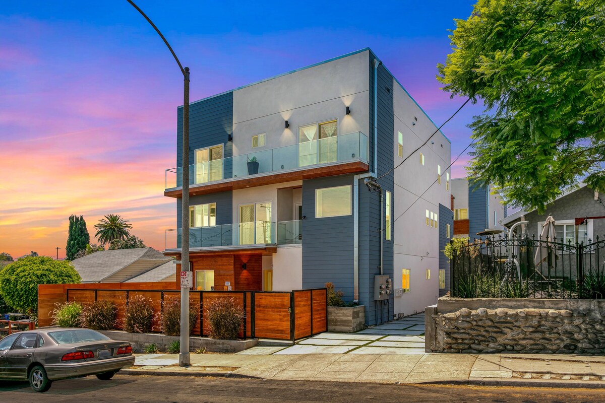 The modern townhome is showcased with a sleek architectural design, featuring multiple levels, large windows, and a balcony. A landscaped front yard with decorative plants surrounds the property, while a driveway provides access to the home. A vibrant sunset adds a warm glow to the scene.