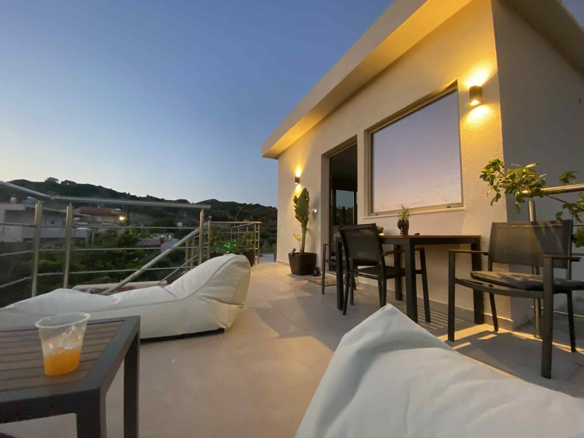 A spacious terrace features comfortable seating options, including bean bag chairs and a dining table. Soft lighting illuminates the area, creating a welcoming ambiance. A scenic view of the surrounding landscape is visible through the large window, enhancing the outdoor living experience.