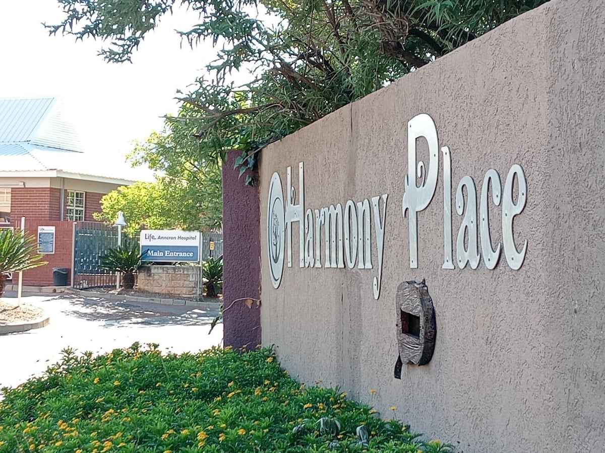 The entrance of Harmony Place is marked by a textured wall with the name prominently displayed in metal lettering. Lush green plants and yellow flowering shrubs surround the base, while a glimpse of a building and signage indicating hospital access is visible in the background.