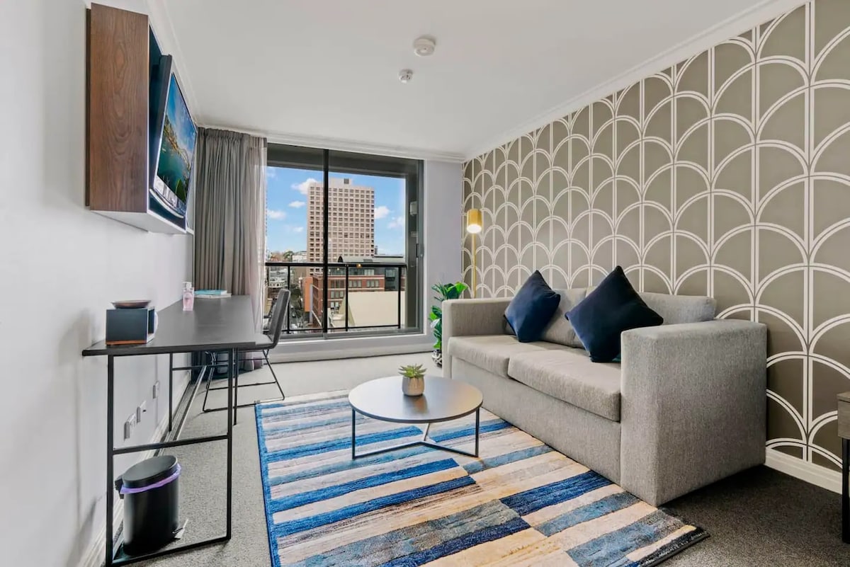 A modern living area features a gray sofa adorned with dark cushions, complemented by a circular coffee table positioned on a striped rug. A sleek desk sits beside a window, providing city views, while patterned wallpaper adds a stylish touch to the space.