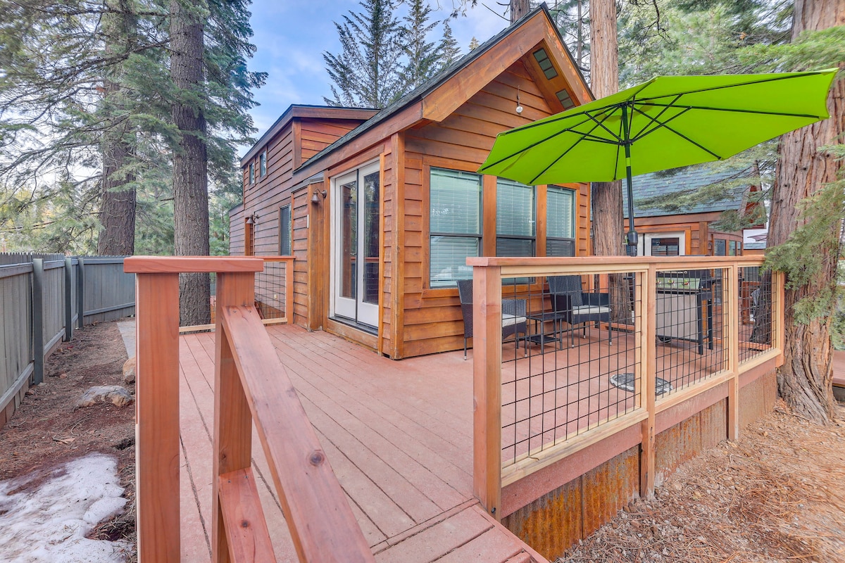A wooden deck is featured, surrounded by tall pine trees, providing a tranquil entrance to the cabin. An umbrella offers shade over a small outdoor seating area with chairs, while large glass doors connect to the interior of the cabin.