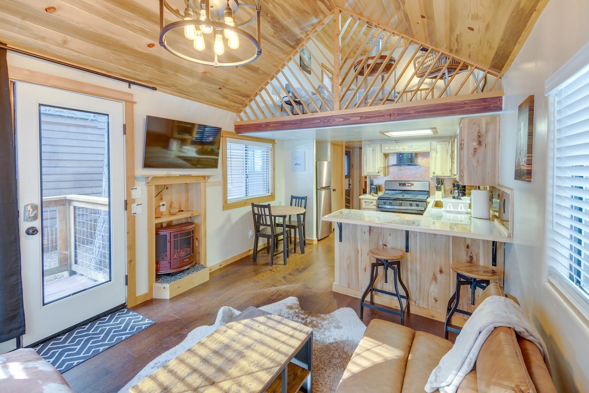 An inviting living space features a comfortable sofa and a mounted flat-screen TV above a stylish electric fireplace. A kitchenette with modern appliances is visible, along with a dining area equipped with two chairs. Natural wood accents enhance the cabin's rustic charm.