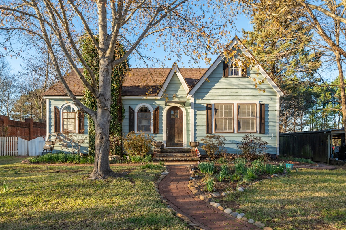 The charming cottage features a light blue exterior with arched windows and brown shutters. A landscaped front yard is bordered by a stone walkway leading to the front door, surrounded by trees and shrubs, providing an inviting approach to the home.