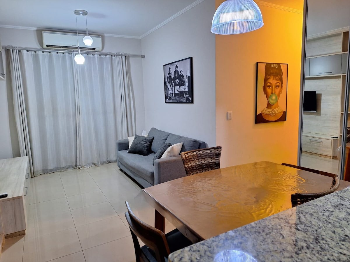 The living area features a comfortable grey sofa accompanied by coordinating pillows, situated under modern pendant lighting. A dining table with wooden chairs is visible, along with light-colored walls and glass sliding doors that brighten the space with natural light.