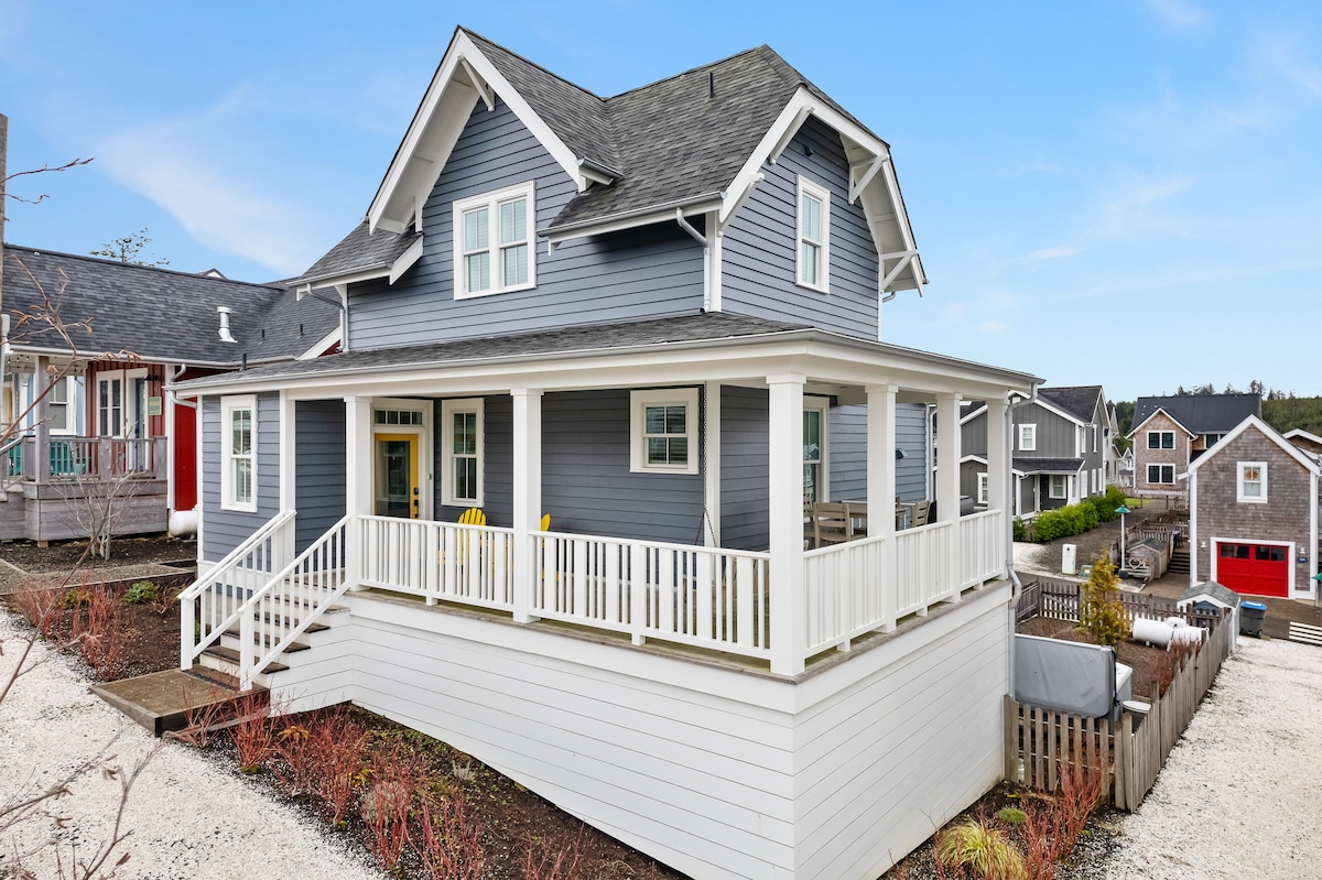 The two-story retreat features a light blue exterior with white trim and a spacious covered front porch. A set of yellow chairs is positioned to create a welcoming seating area, while the landscaped yard includes clean pathways and nearby homes visible in the background.