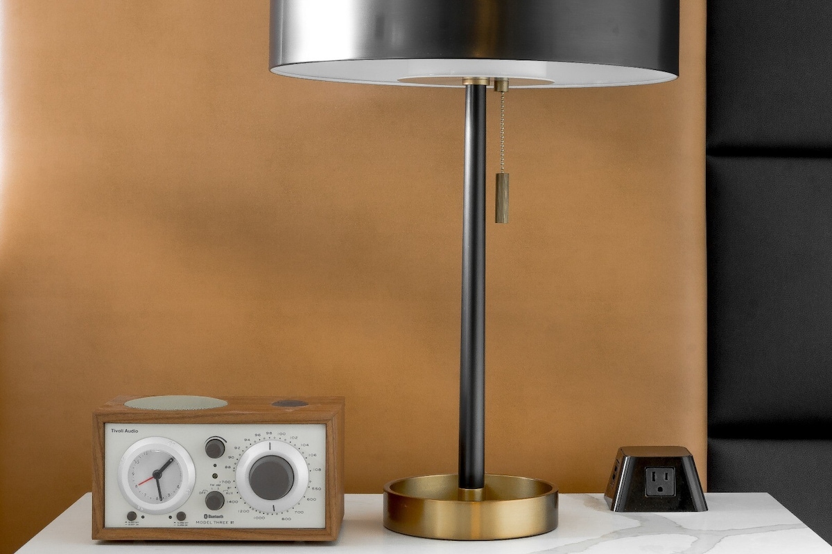 A bedside table is presented with a modern lamp featuring a metal base and a pull chain. Next to it, a wooden radio with dials and a power outlet adds functionality, complementing the warm tones of the background wall.