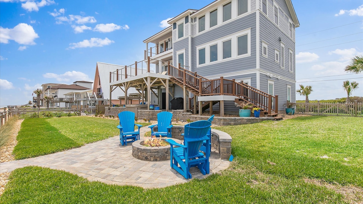 A spacious backyard is showcased, featuring a circular stone patio with bright blue Adirondack chairs arranged around a fire pit. The modern multi-level home with large decks overlooks the well-manicured lawn and is surrounded by palm trees, contributing to a relaxed outdoor atmosphere.