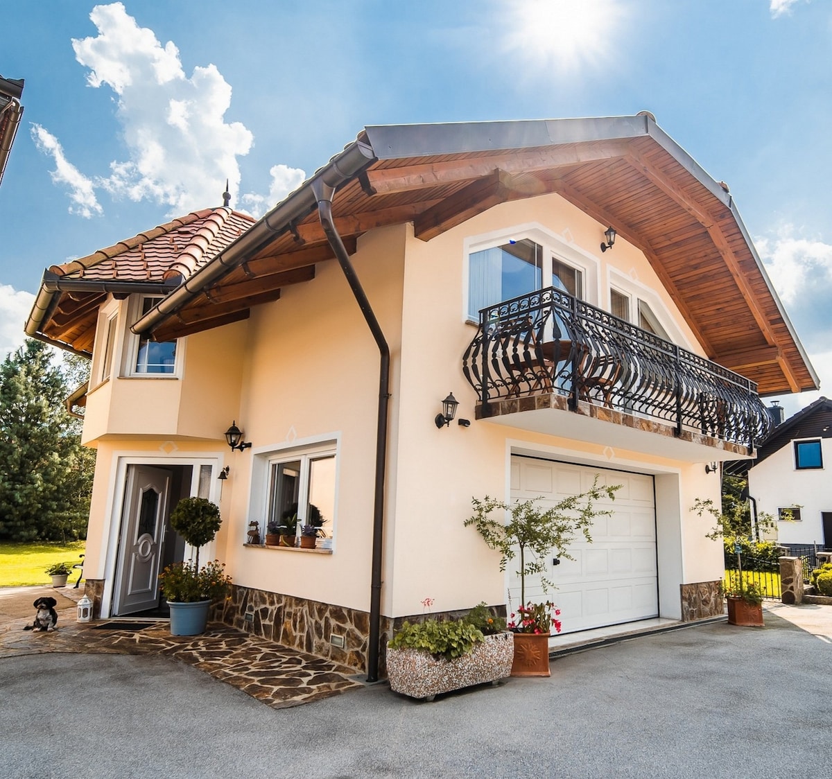 A two-story house features a charming balcony with decorative railings and a garage at the front. Sunlight gleams on the light-colored exterior, surrounded by greenery. Potted plants are positioned near the entrance, enhancing the inviting appearance of the home.