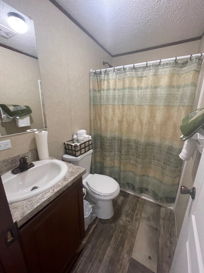 The compact bathroom includes a sink with a mirrored cabinet above and a shower area enclosed by a patterned curtain. Fresh towels and toilet paper are neatly arranged, with a small basket containing additional toiletries also visible.