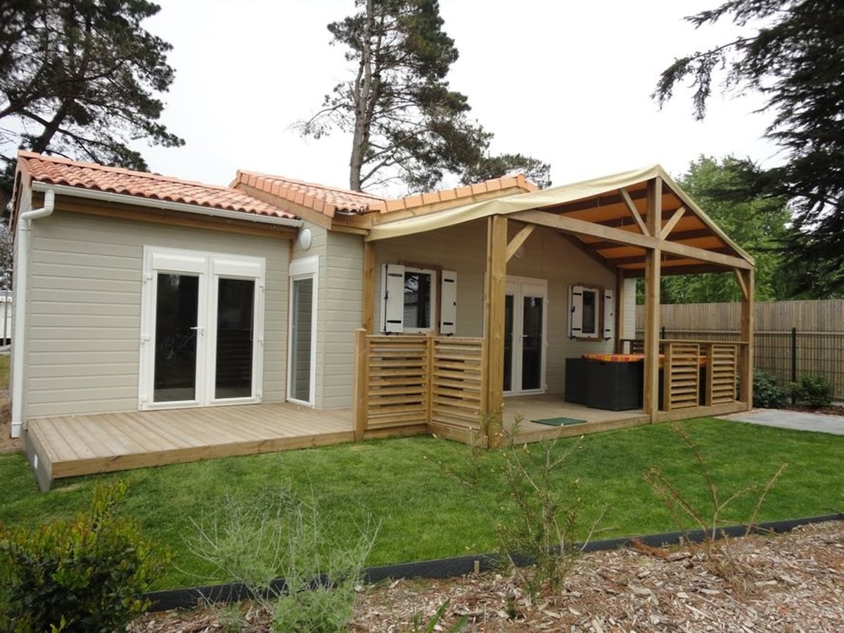 A modern chalet is showcased from an exterior angle, featuring a covered terrace with wooden flooring. The structure is surrounded by green grass and landscaping, with large windows allowing for natural light. The roof has a warm tile design, enhancing the overall aesthetic.
