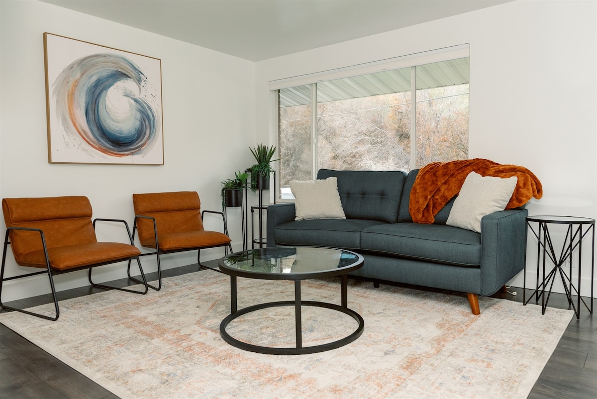 A well-arranged living room features a comfortable blue sofa adorned with a textured orange throw, seated opposite two stylish armchairs. A round glass coffee table sits on a soft area rug, with natural light filtering in through a large window revealing a serene outdoor view.