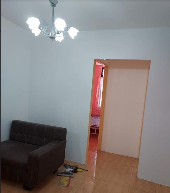 A small, well-lit room features a comfortable dark brown armchair positioned against a white wall. A simple chandelier with five bulbs hangs from the ceiling. A doorway leads to another room, partially visible, showcasing a bed with light-colored bedding.