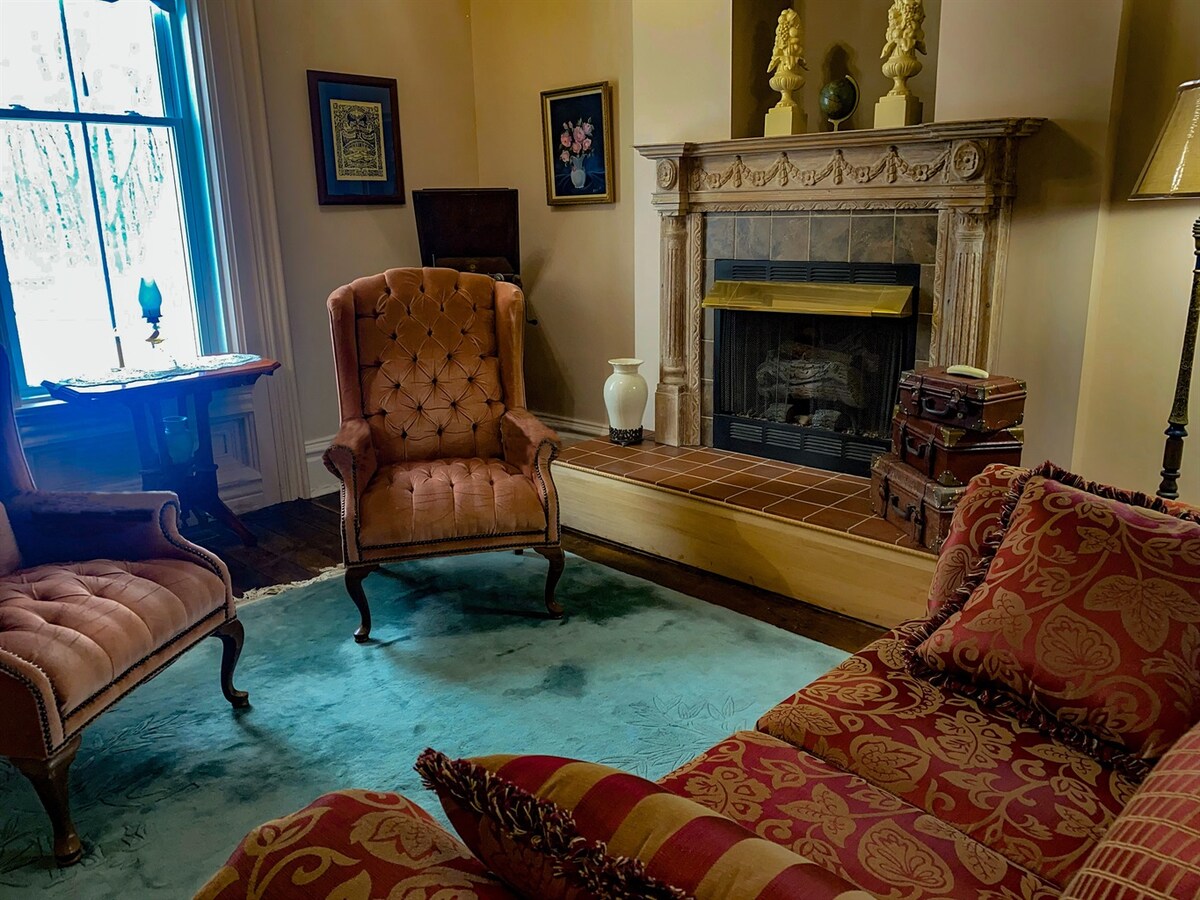 A cozy living room is presented, featuring a large fireplace with a detailed mantle. Two tufted armchairs face a vintage sofa, all arranged on a textured area rug. Large windows allow natural light to illuminate the space, highlighting the antique decor.