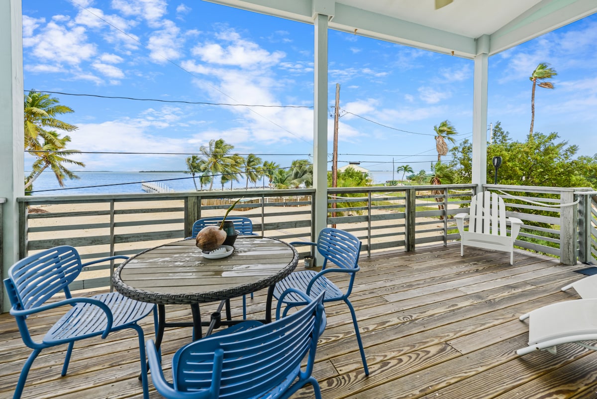 A covered deck area features a round table with six blue chairs, offering views of the water and surrounding palm trees. The space is illuminated by natural light, creating a relaxing spot to enjoy the outdoors.