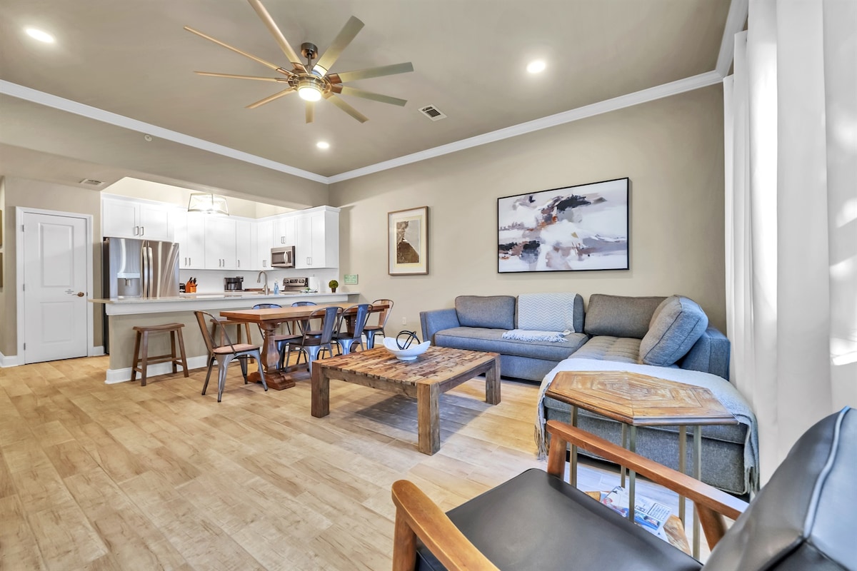 An open-concept living area features a light wood floor complemented by a large coffee table and a comfortable sofa. Bright natural light fills the room through large windows, and a ceiling fan provides airflow. A kitchen island with seating enhances the communal space.