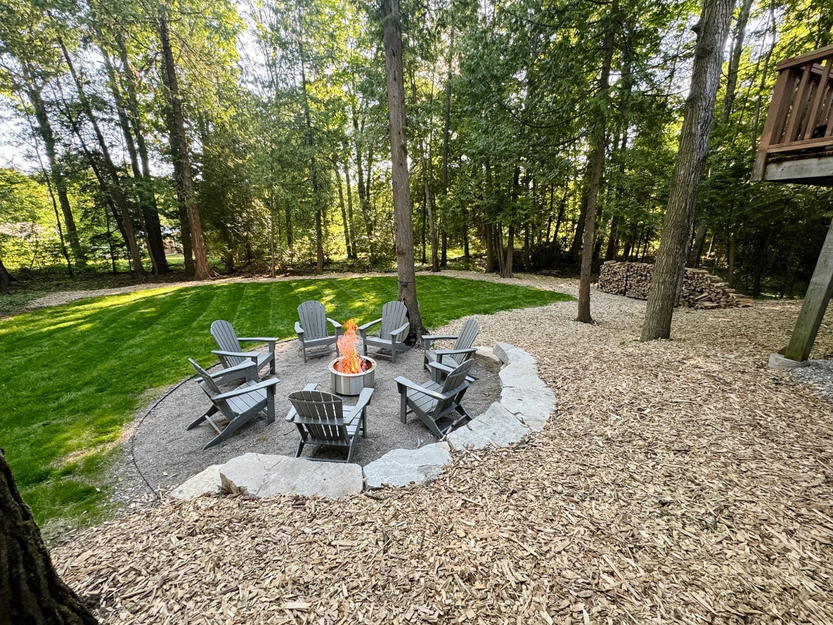 A circular firepit area is surrounded by multiple Adirondack chairs, set on a gravel base. Tall trees provide a natural backdrop, and a neatly landscaped lawn adds to the serene atmosphere. Stacked wood is visible nearby, suggesting a cozy gathering space.