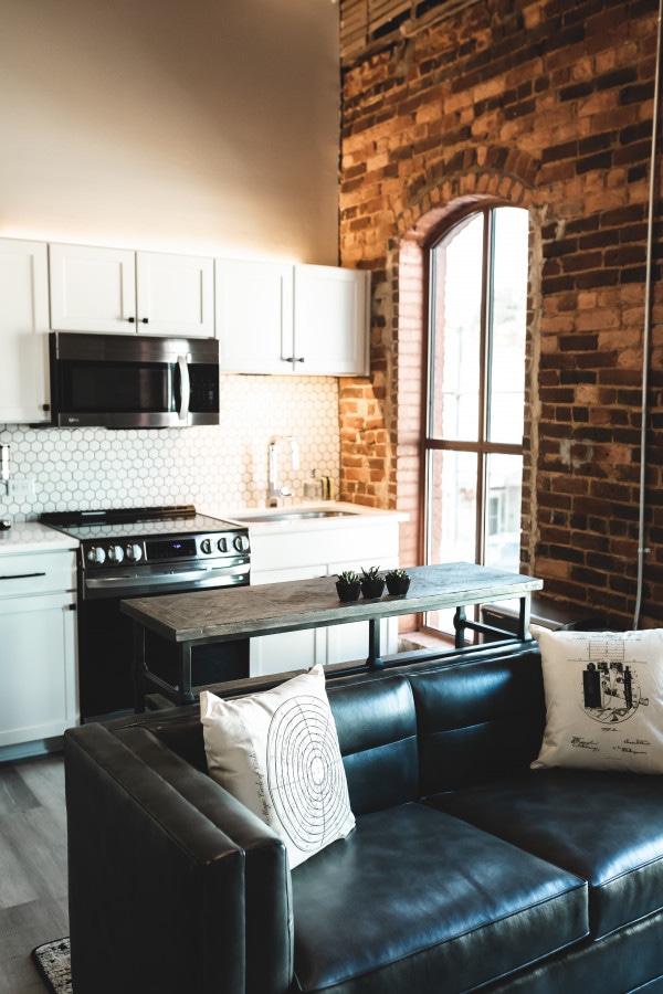 The kitchen area features modern white cabinetry and a stainless steel appliance suite, complemented by exposed brick walls. A sleek, black leather sofa is placed in the foreground, accompanied by decorative pillows and a wooden coffee table, enhancing the open layout.