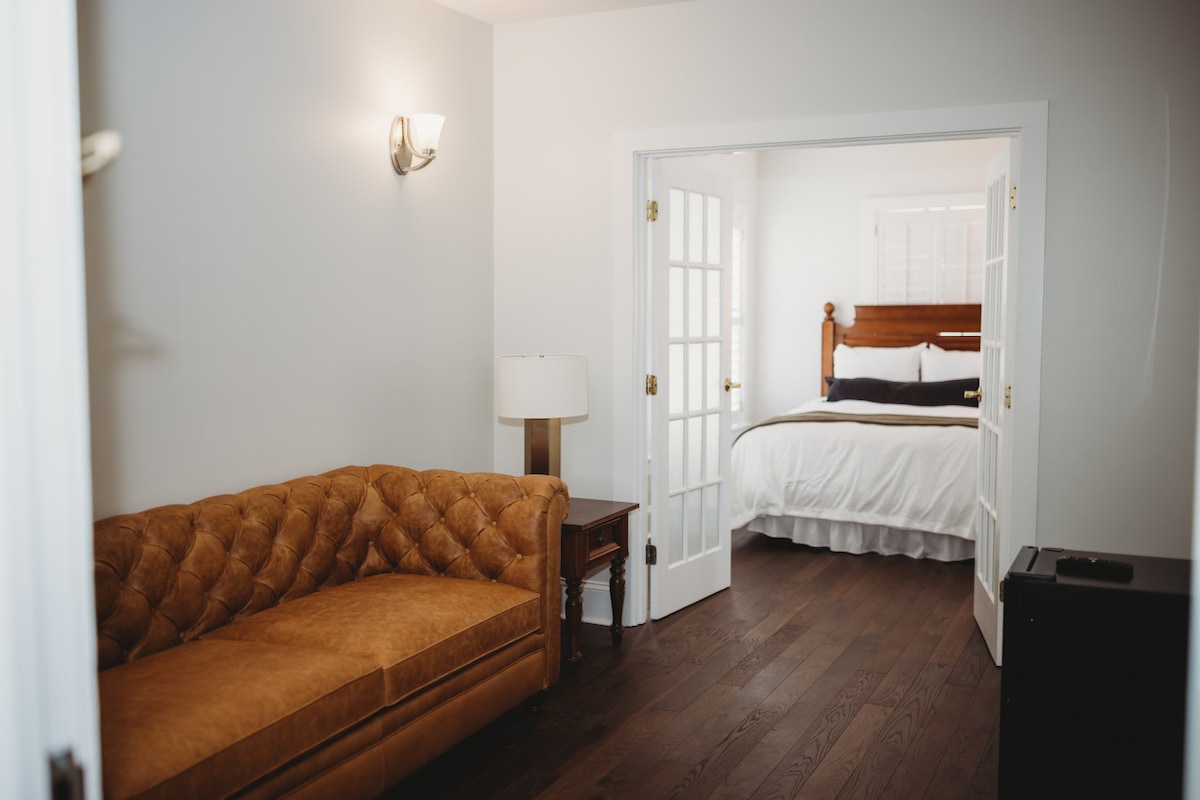 A sitting area is visible, featuring a tufted brown sofa beside a wooden side table and lamp. Covered french doors lead to the bedroom, which contains a spacious bed with white linens. Natural light filters in through a window, highlighting the warm wooden flooring.