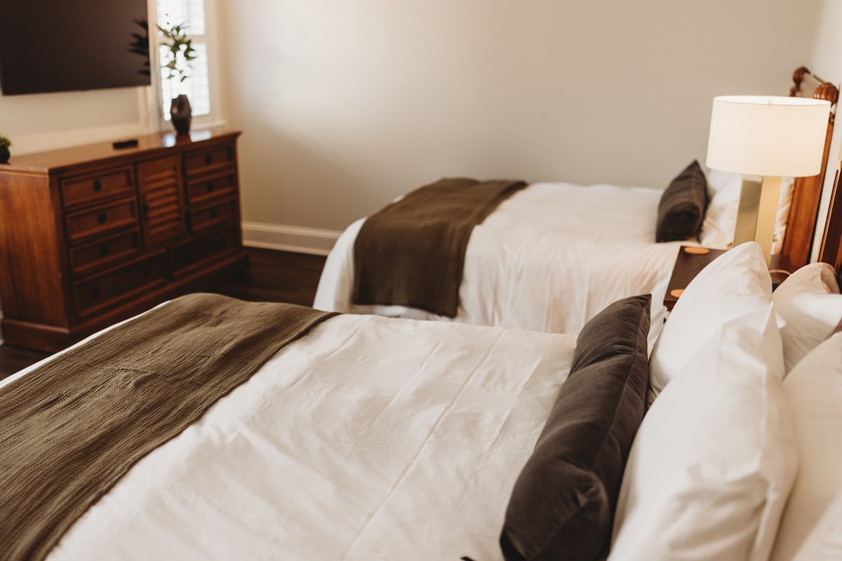 The room features two spacious queen beds dressed in luxurious white linens, complemented by dark throw blankets. A wooden dresser is situated against the wall, with a television mounted above. Natural light enters through a nearby window, creating a bright and inviting space.