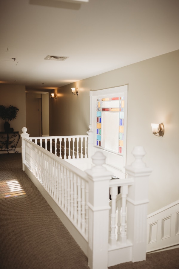 A well-lit hallway features a white banister along the upper railing. Natural light filters through a decorative stained-glass window, adding color to the neutral-toned walls. Soft glowing sconces accent the space, contributing to a serene atmosphere.